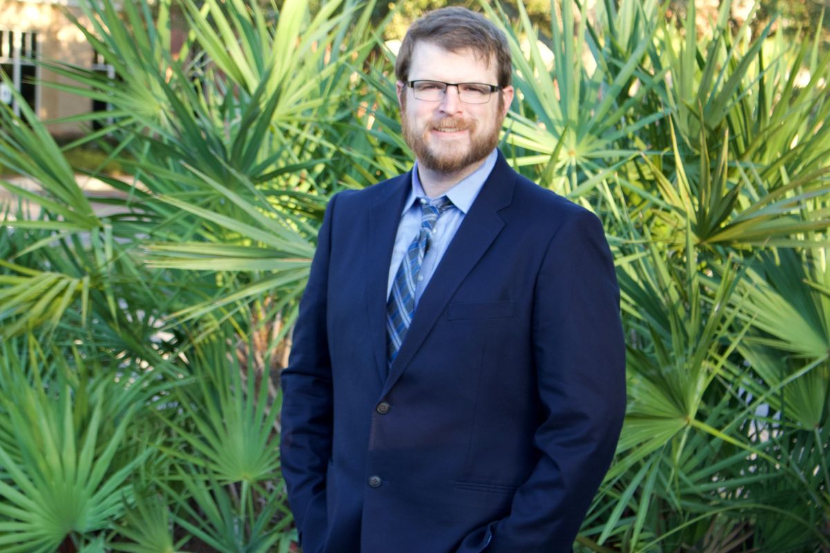 Samuel Price in a navy suit and a blue tie, standing in front of palmettos